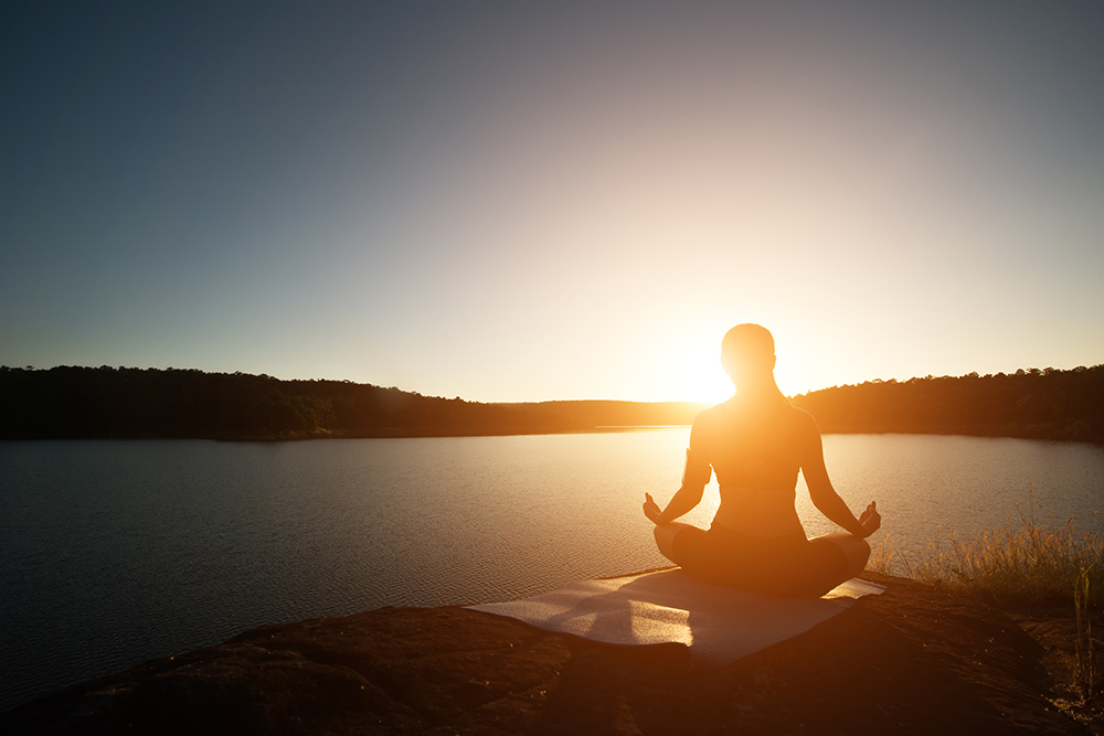 Silhouette of healthy woman is practicing yoga at mountain lake during sunset.